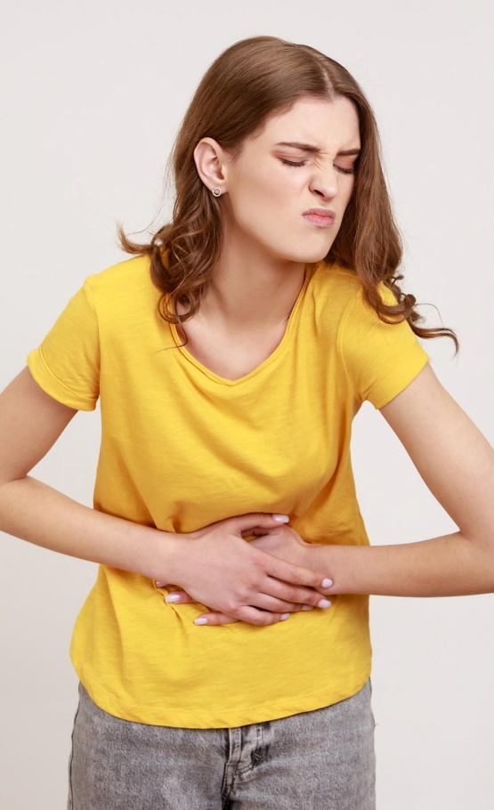 portrait of unhappy sick teenager girl with brown hair in casual style tshirt standing holding her belly with hands stomach cramps or period pain indoor studio shot isolated on gray background