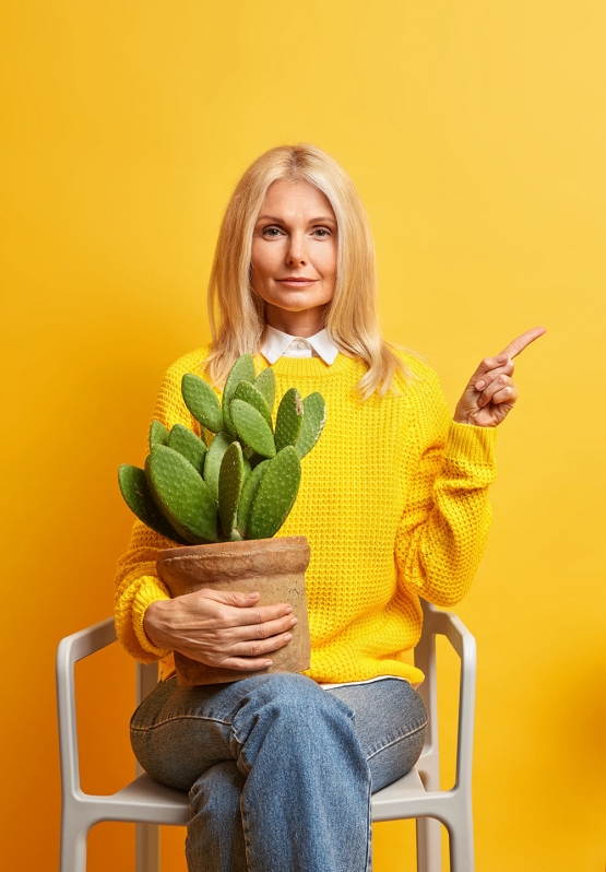 serious middle aged woman poses with cactus at chair looks confidently and points away on copy space