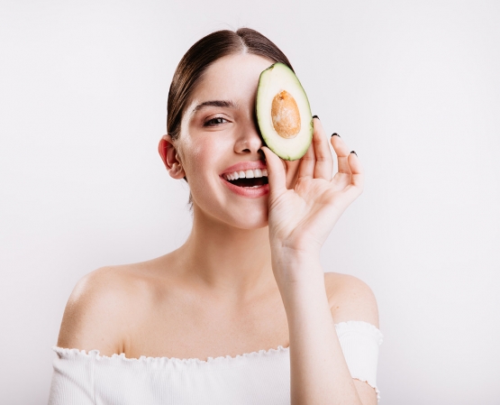 woman without makeup with clean skin is smiling posing with slice of avocado for portrait on white wall